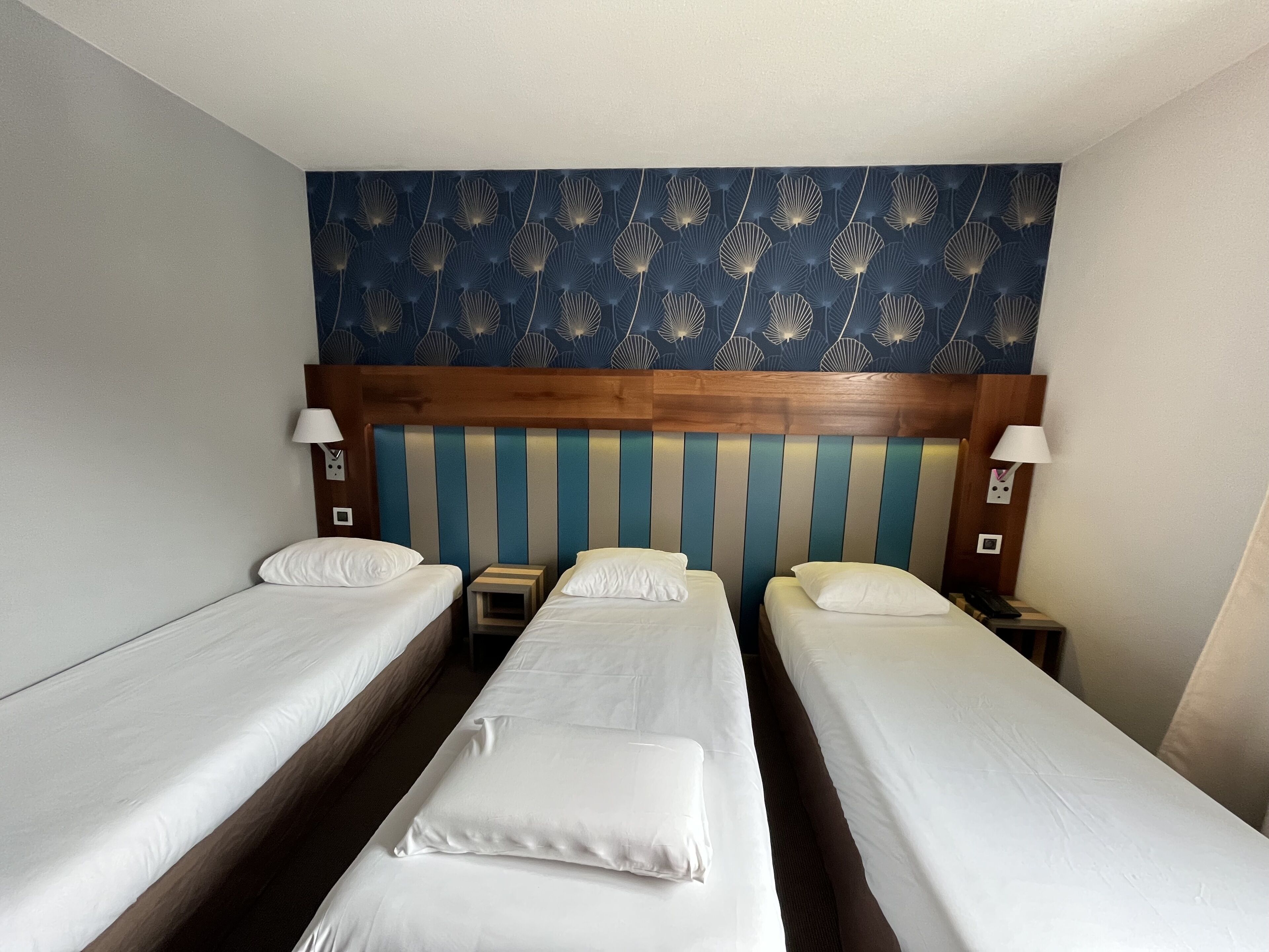 Quarto de hotel com três camas de solteiro, cabeceira de madeira e um padrão decorativo na parede.