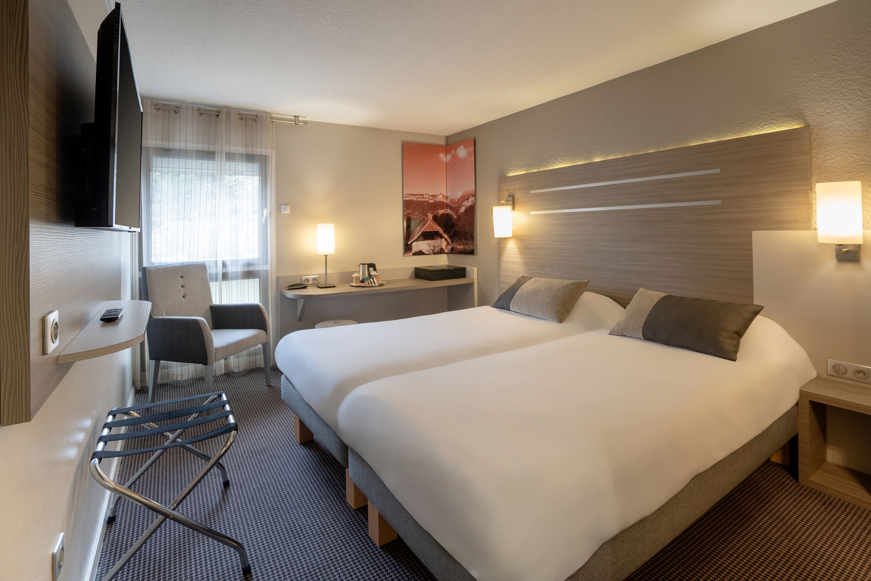 Une chambre d’hôtel avec deux lits, un bureau, une chaise, une télévision et une lampe fixée au mur.