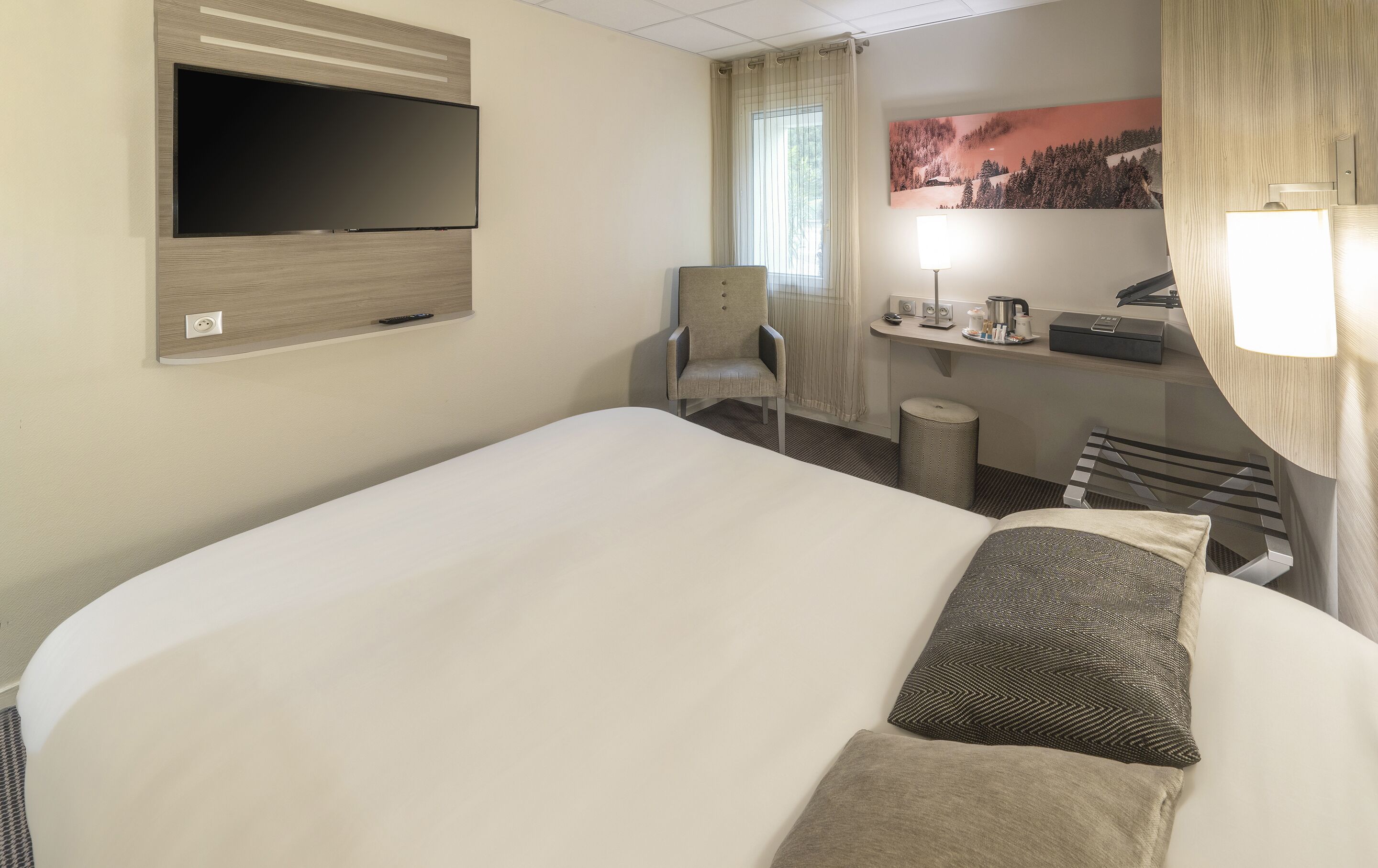 A hotel room with a large bed, a TV mounted on the wall, a chair, a desk with a lamp, and a painting on the wall.