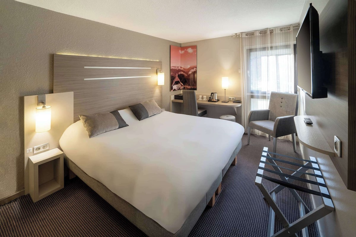 Une chambre d’hôtel avec un grand lit, des tables de chevet avec des lampes, un bureau avec une chaise, une télévision et une fenêtre avec des rideaux.