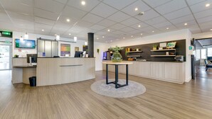 Un hall d’hôtel moderne avec un comptoir d’accueil, une table ronde ornée d’un vase et un grand écran diffusant un paysage.