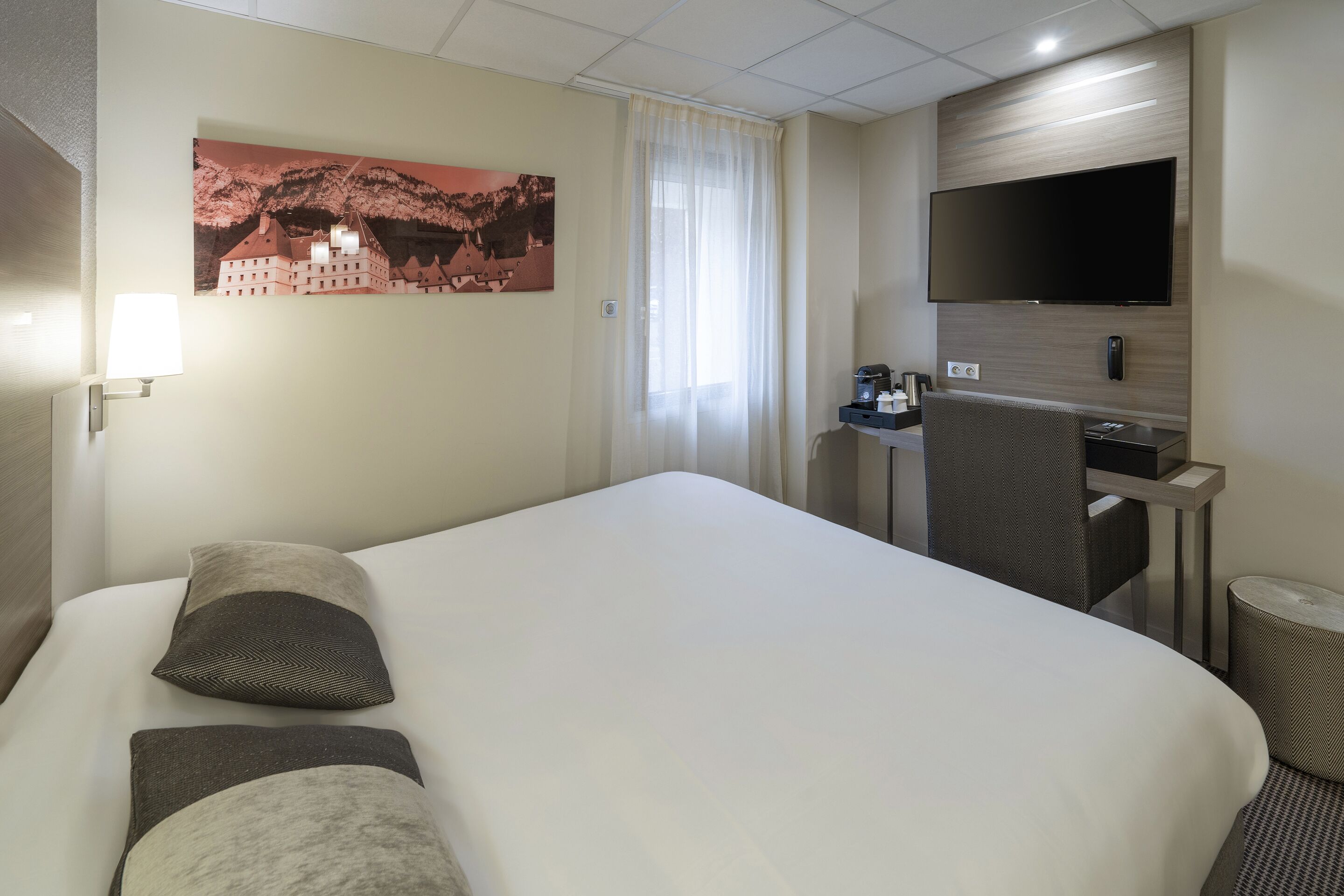 A hotel room with a large bed, a flat-screen TV mounted on the wall, a desk with a chair, and a painting of a castle on the wall.