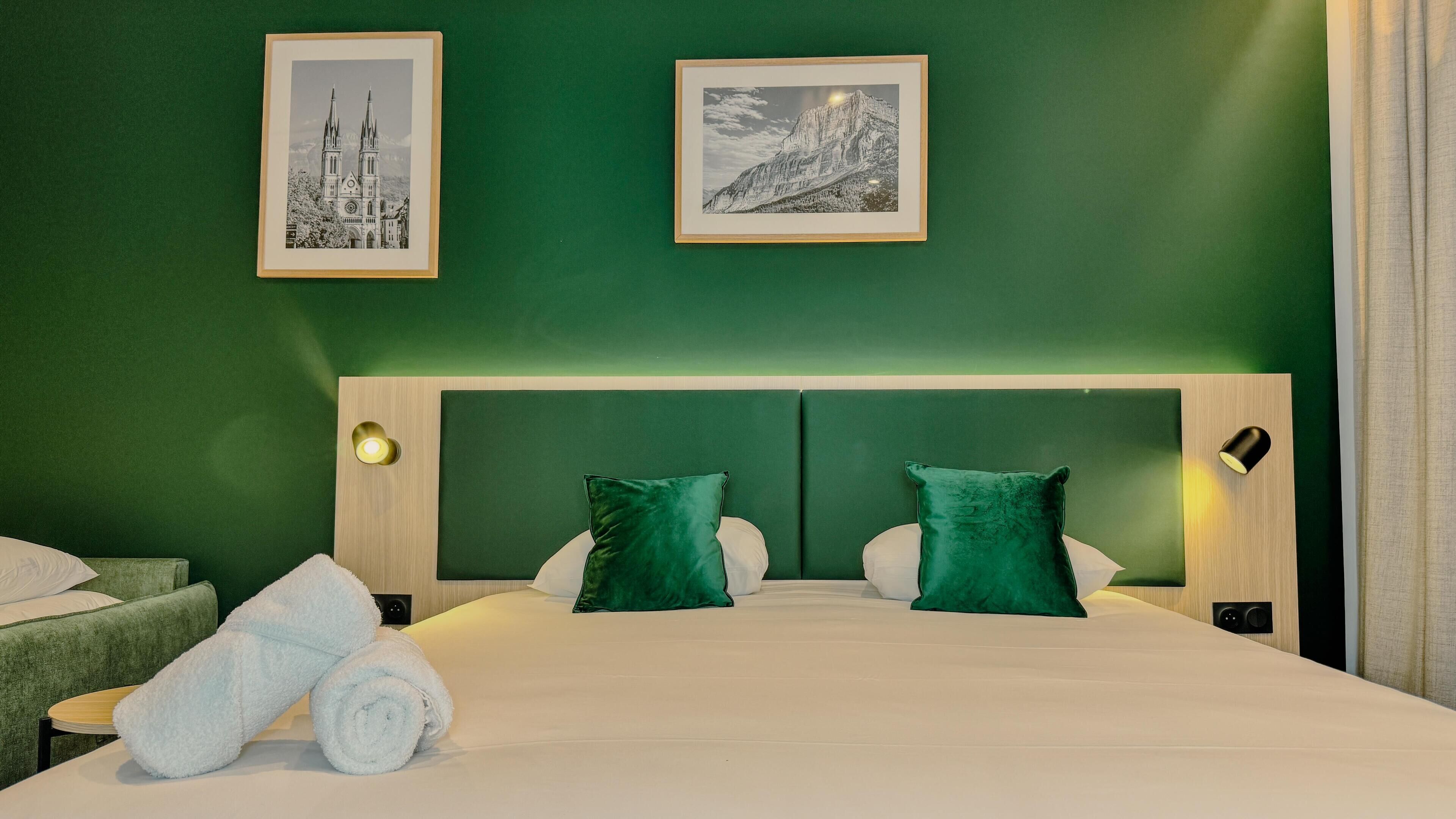 Une chambre d’hôtel avec un mur vert, un grand lit avec une literie blanche, deux oreillers verts et deux tableaux encadrés au-dessus du lit.