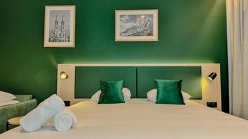 Une chambre d’hôtel avec un mur vert, un grand lit avec une literie blanche, deux oreillers verts et deux tableaux encadrés au-dessus du lit.