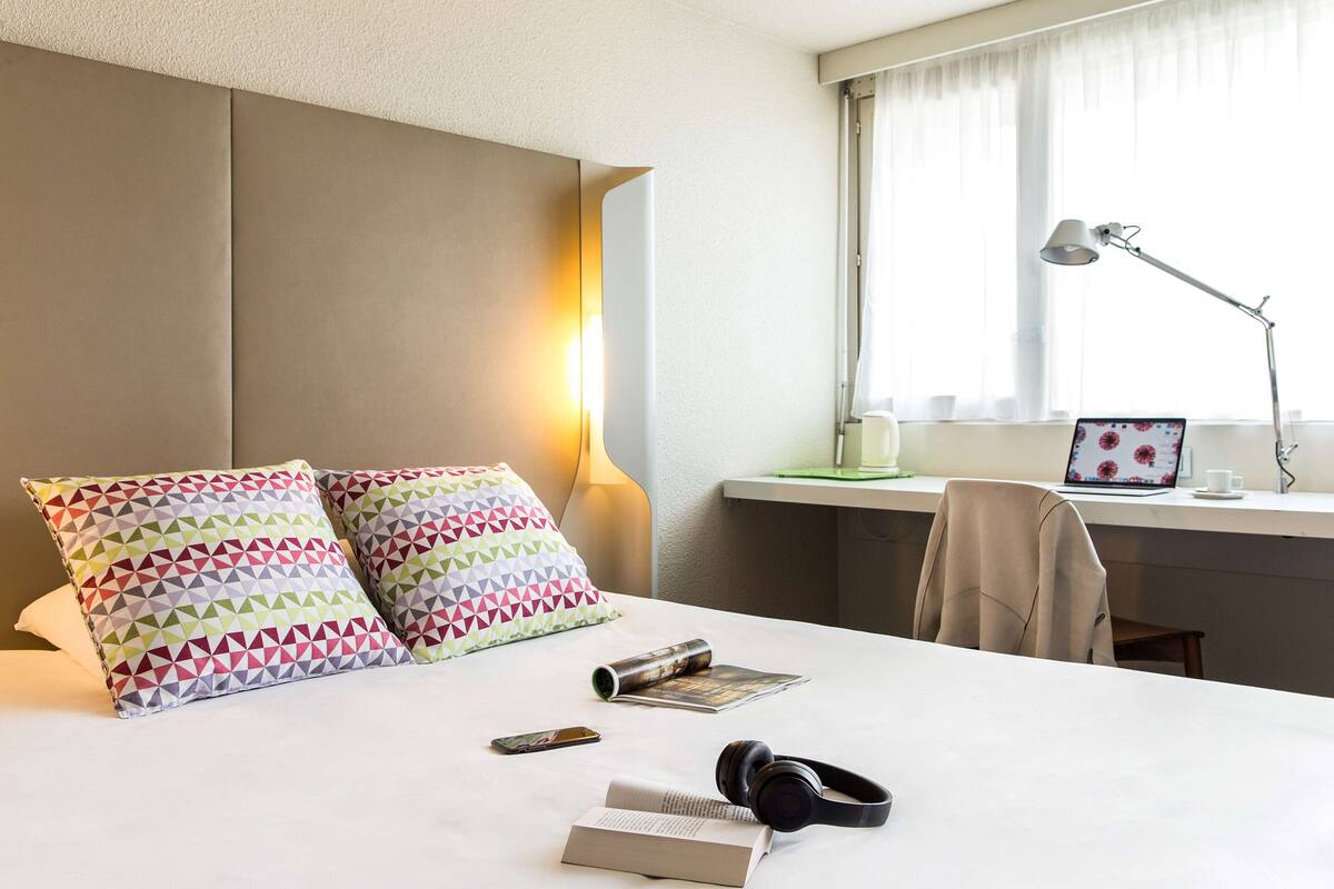 Une chambre d’hôtel avec un lit, des oreillers, un bureau avec un ordinateur portable, une lampe et une chaise.