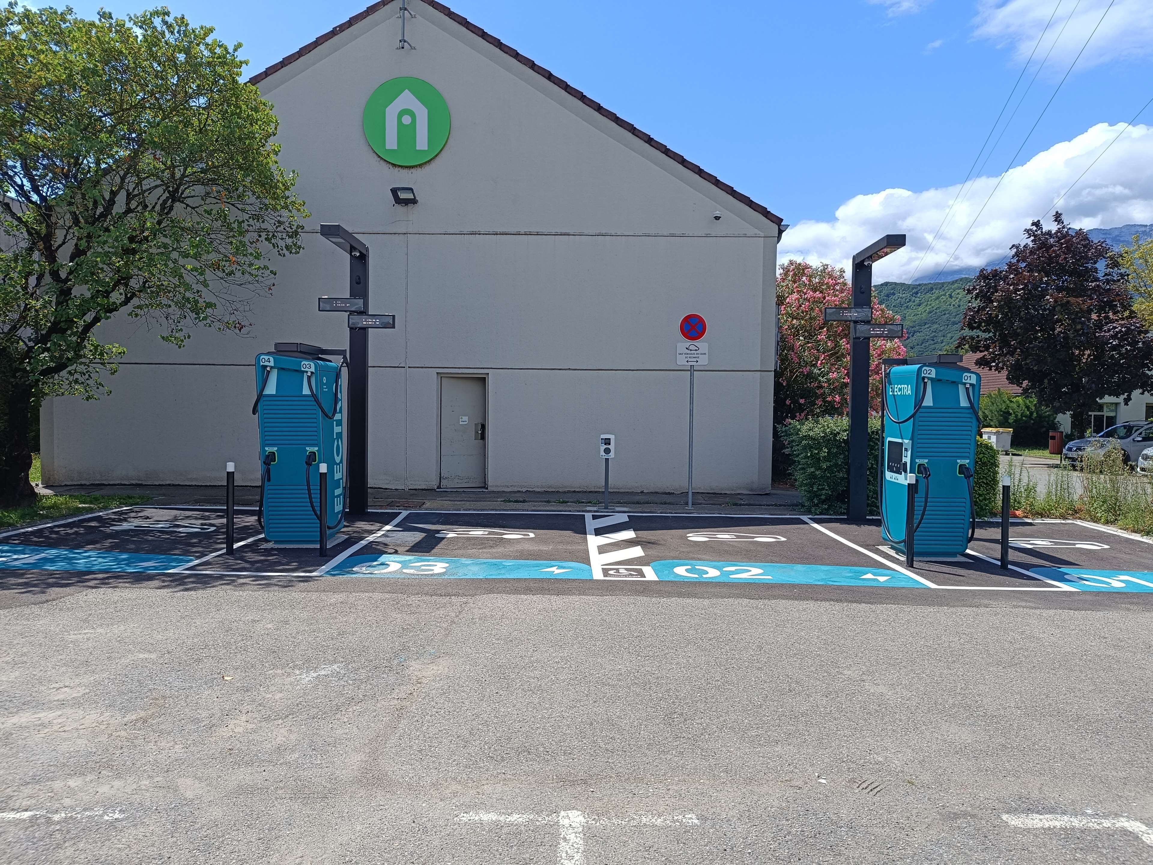 Un edificio con un logotipo verde, dos estaciones de carga para vehículos eléctricos y un estacionamiento con marcas azules.