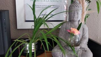 Una estatua gris con un florero de flores sobre un estante de madera, una planta en maceta y un cartel enmarcado en la pared.