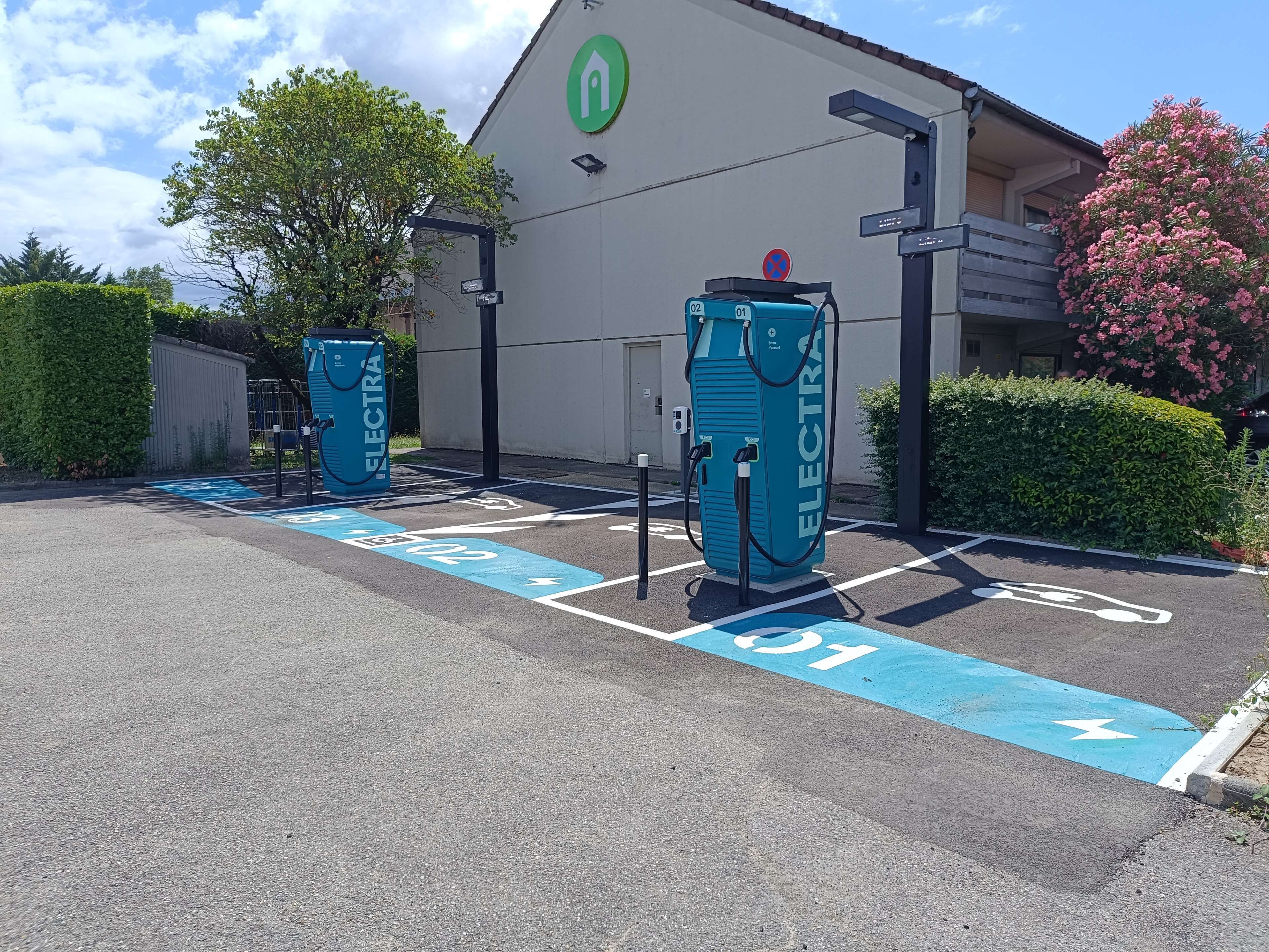 Une borne de recharge pour véhicules électriques dotée de deux points de recharge, de places de stationnement délimitées et d’un bâtiment arborant un logo vert.