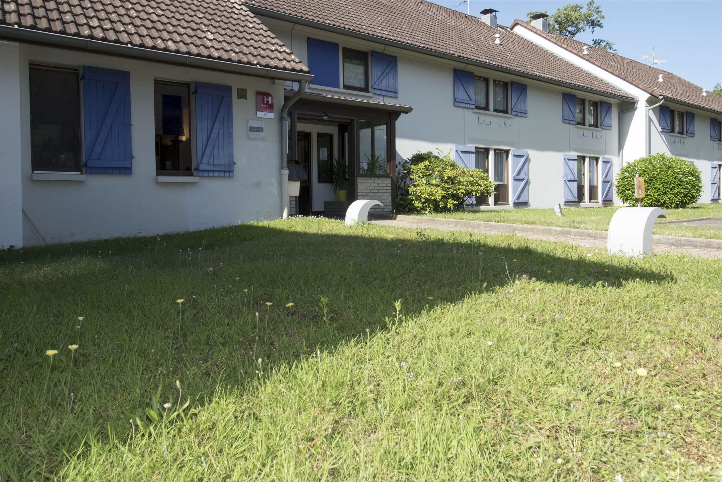 A building with a tiled roof, multiple windows with blue shutters, a small entrance with a door, and a well-maintained lawn with grass and small plants.