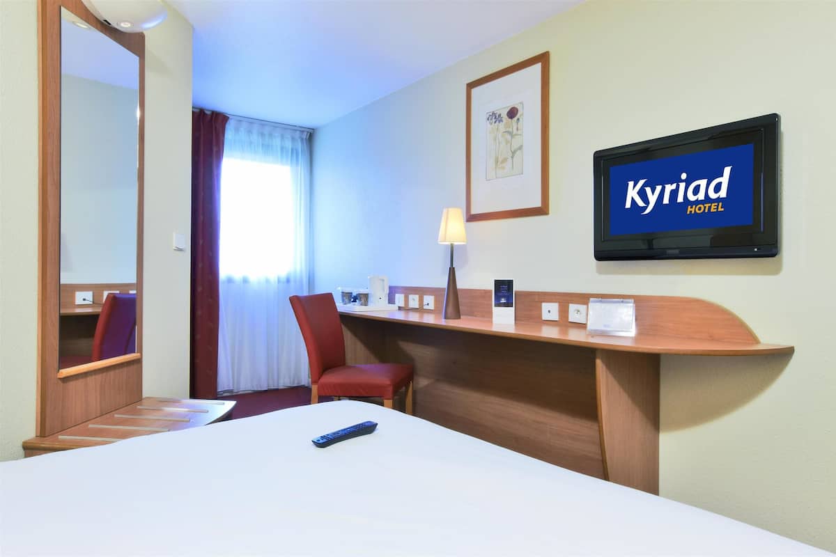 Hotelkamer met een bed, bureau, stoel, televisie waarop 'Kyriad Hotel' staat, en een raam met gordijnen.