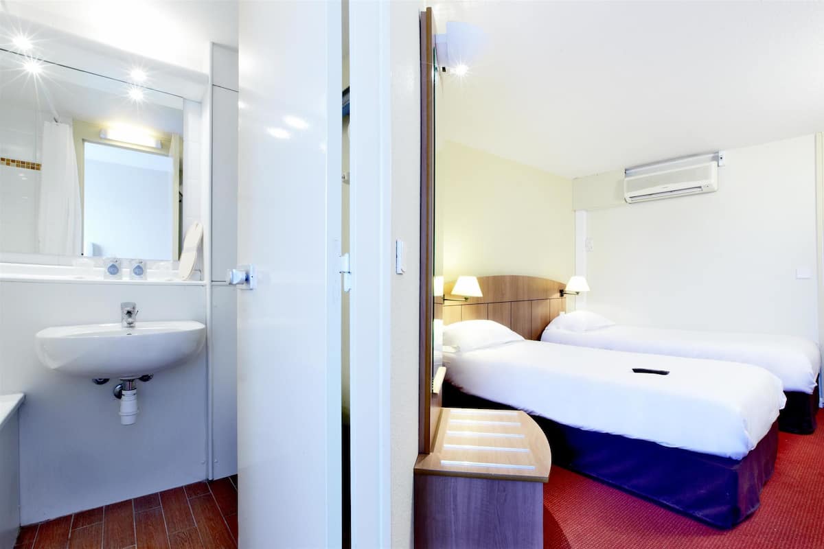 Hotelkamer met een wit wastafel, een spiegel, twee bedden met witte lakens, een nachtkastje en een rood tapijt.
