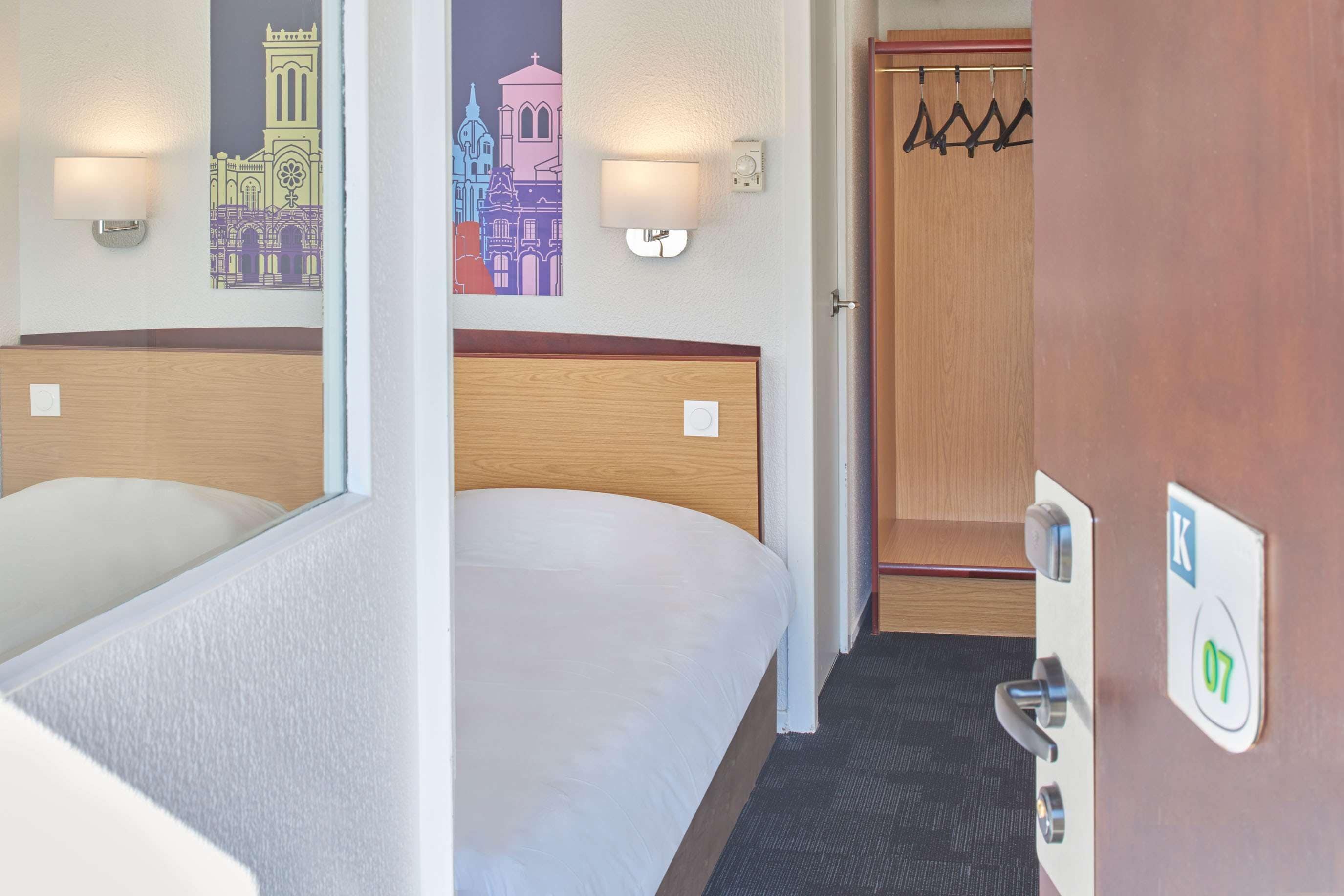 A hotel room with two beds, a wooden headboard, a wall-mounted light, a coat rack, and a door with a keyhole.