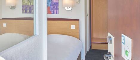 Habitación de hotel con dos camas, cabecero de madera, lámpara de pared, perchero y puerta con cerradura.