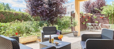 Un área de patio al aire libre con mobiliario de mimbre, una mesa y sillas. Hay plantas en macetas y una valla de madera al fondo.
