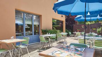 An outdoor patio with tables and chairs, blue umbrellas, and a building with large windows.