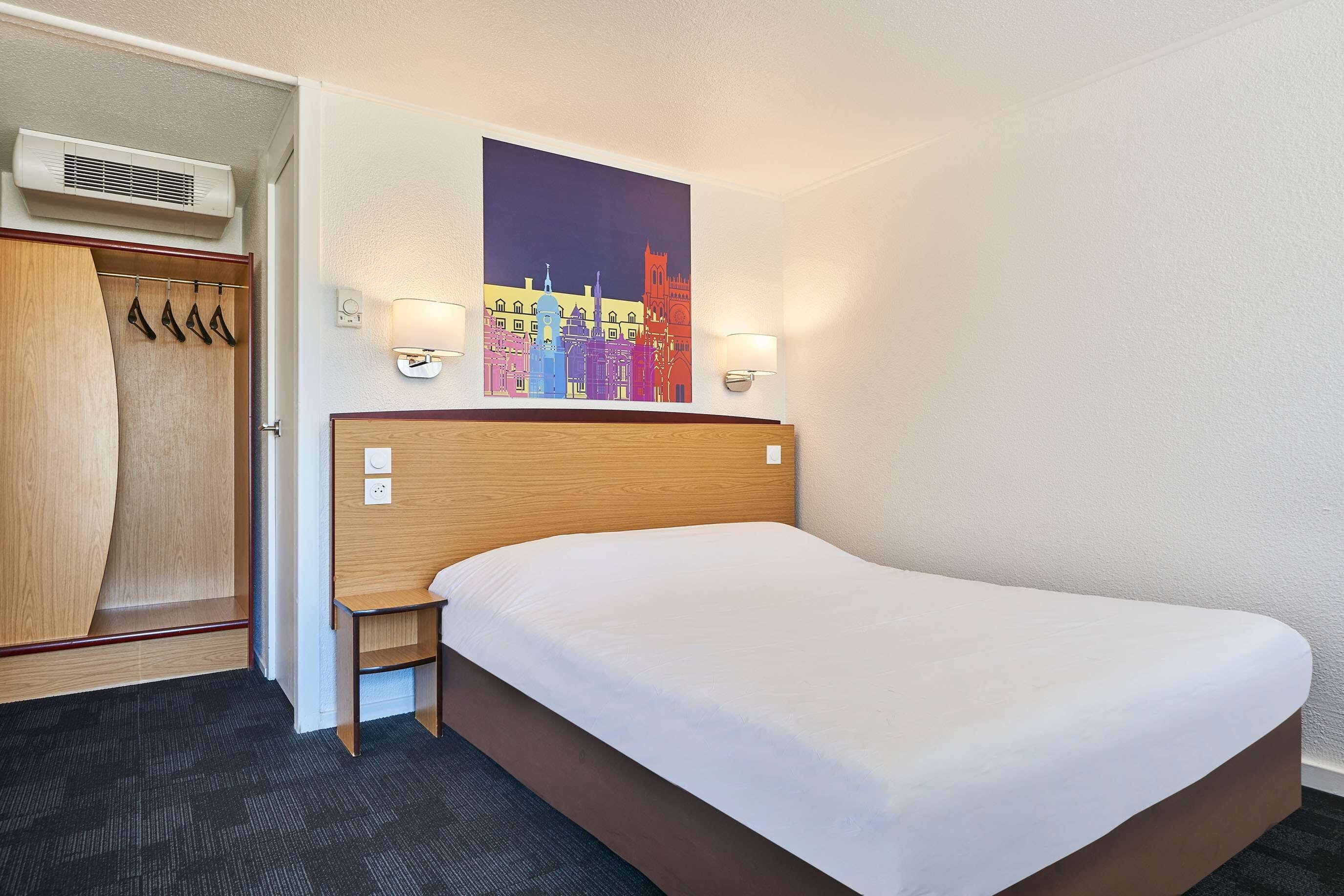A hotel room with a bed, a wardrobe, a coat rack, and a wall-mounted artwork.