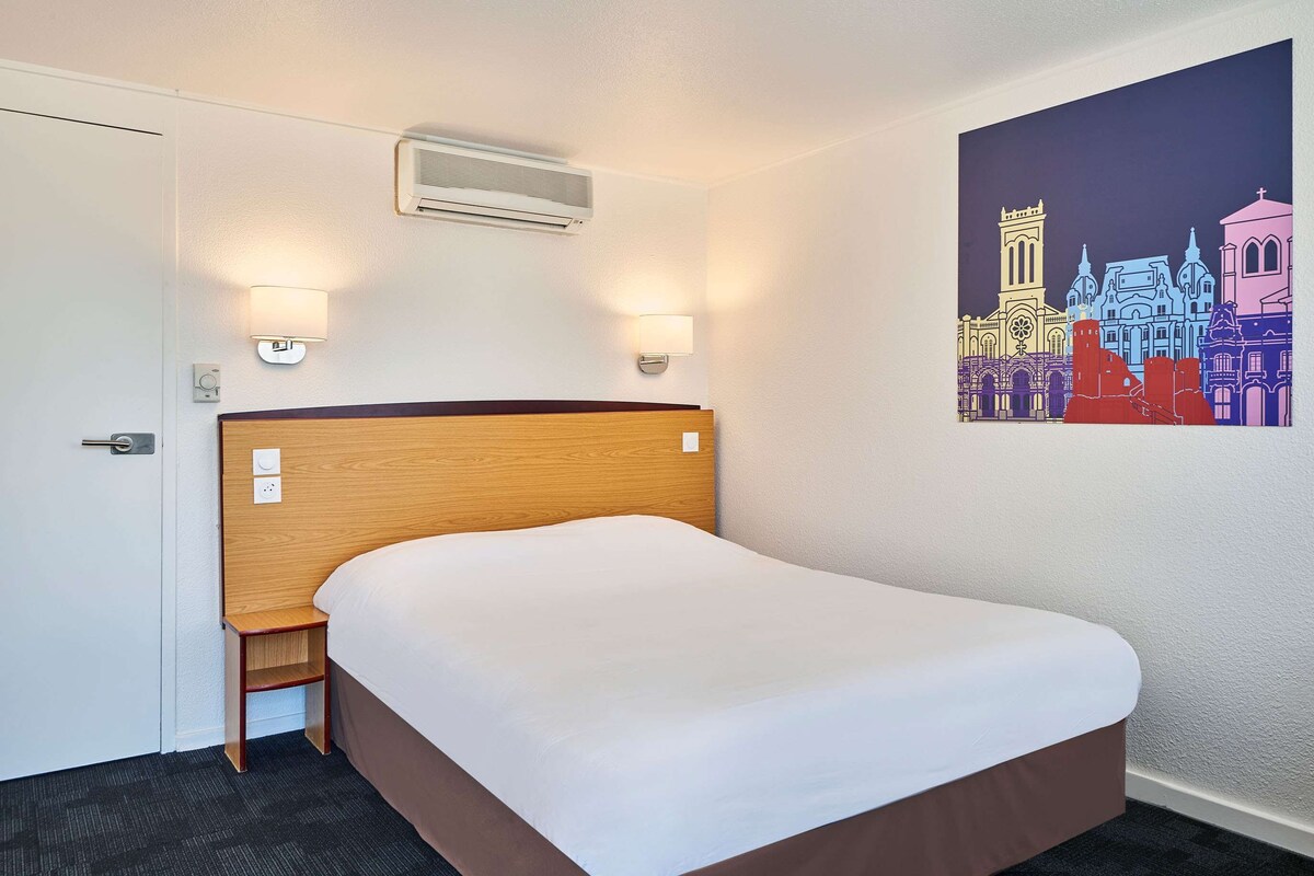 Habitación de hotel con cabecera de madera, una cama individual, dos lámparas fijadas a la pared, un equipo de aire acondicionado y un cuadro de un paisaje urbano colorido en la pared.