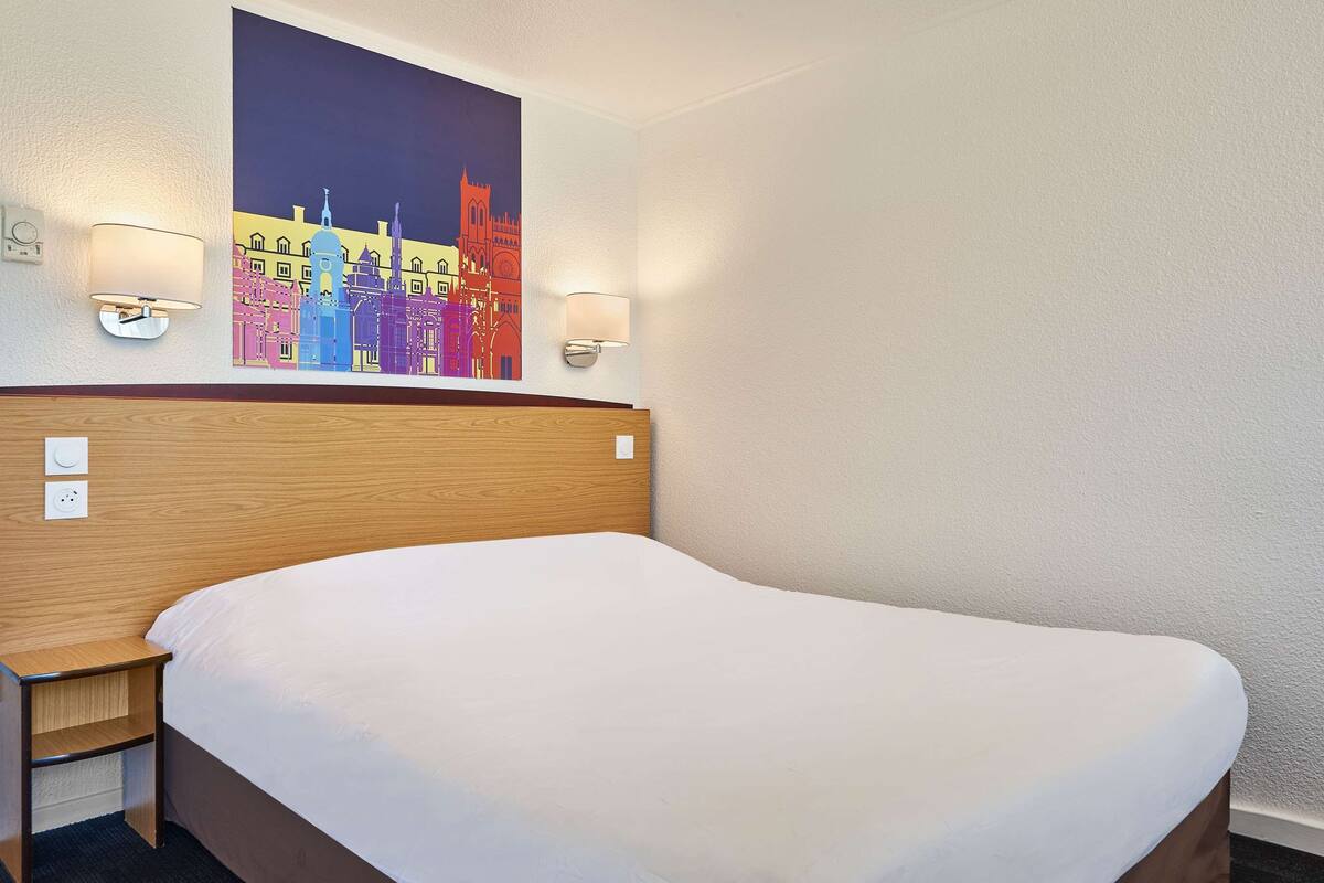 A hotel room with a large bed, a wooden headboard, two wall-mounted lamps, and a colorful cityscape painting above the bed.