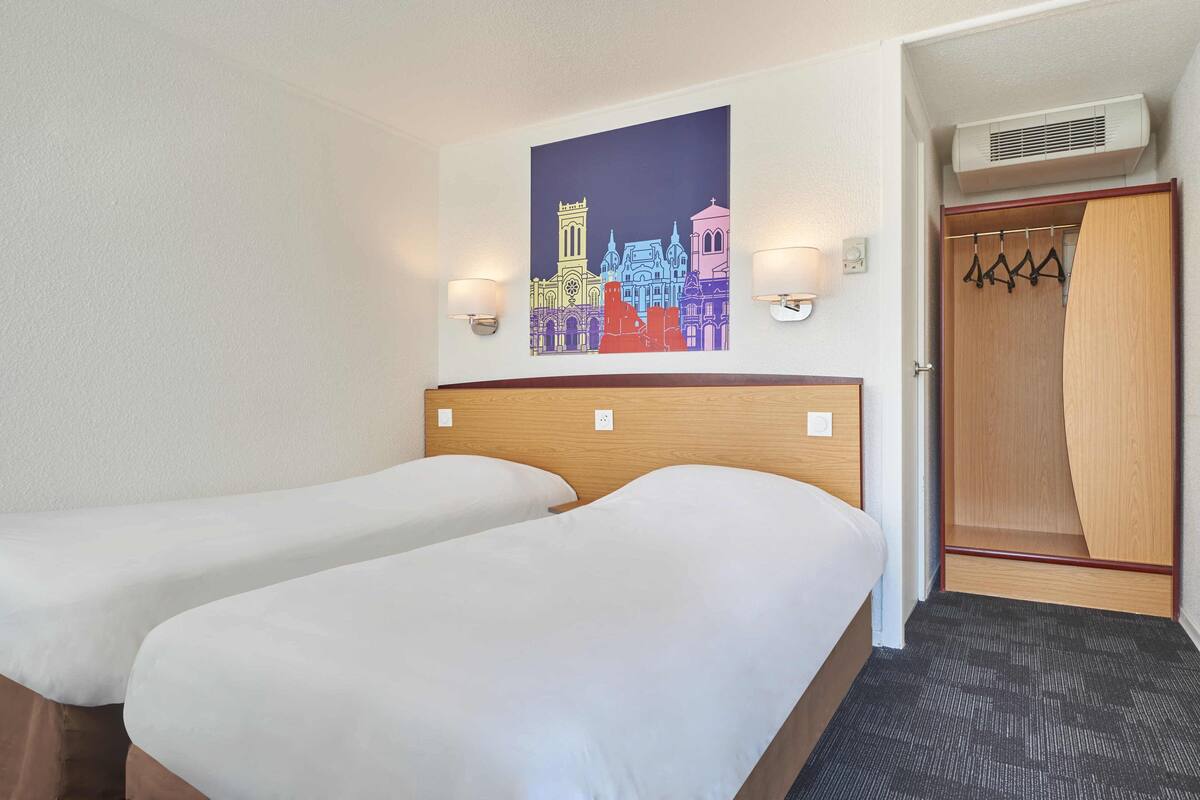 A hotel room with two single beds, a wooden headboard, a wall-mounted painting of a cityscape, and a wooden wardrobe with a mirror.