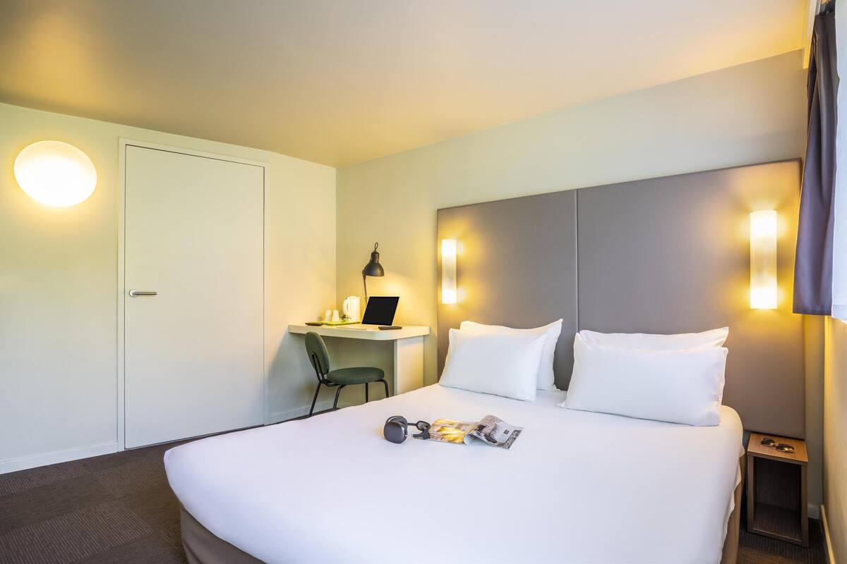 A hotel room with a bed, a desk with a laptop, a chair, and a nightstand with glasses.