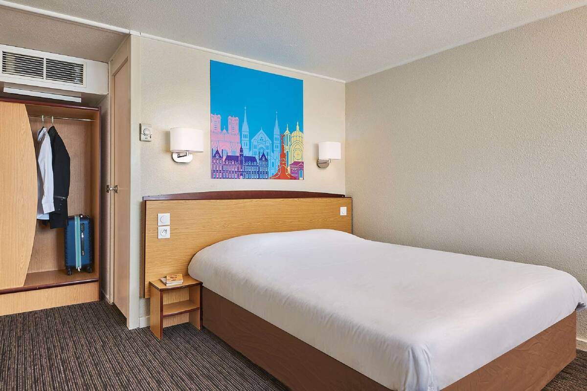 Une chambre d’hôtel avec un grand lit, une armoire, un bureau et un tableau représentant un paysage urbain accroché au mur.