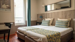 A hotel room with a bed, a desk, a chair, a window with curtains, and a mirror on the wall. - Plazza hôtel (Nîmes)