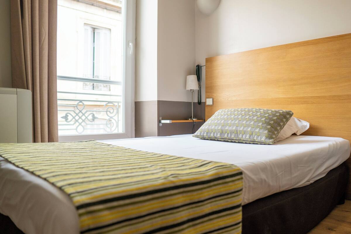 A hotel room with a wooden headboard, a bed with striped bedding, a bedside table with a lamp, and a view of a building outside.