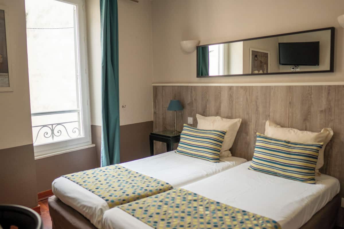 A hotel room with two beds, a television, a window with curtains, and a wall-mounted mirror.