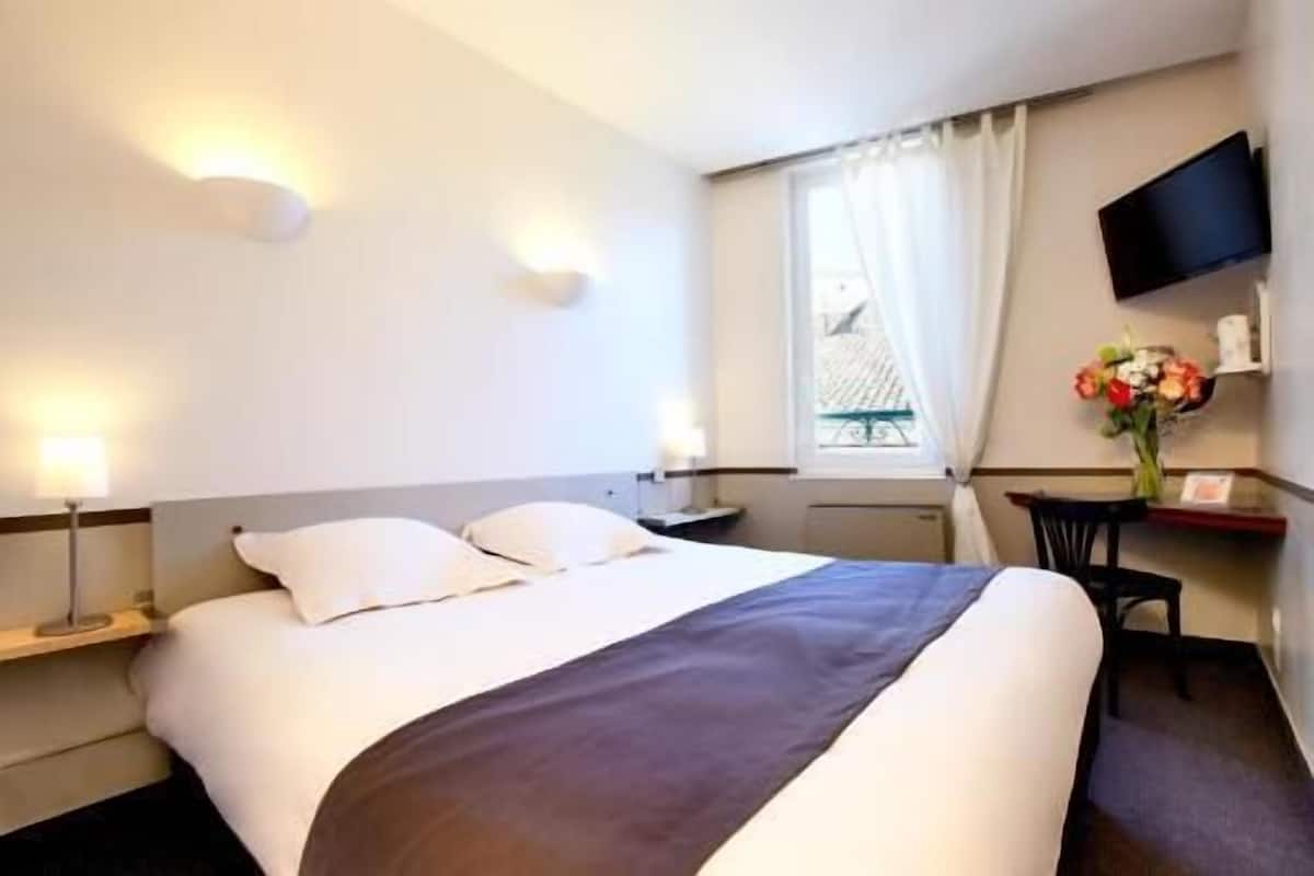 A hotel room with a bed, bedside tables, a desk, a chair, a TV, and a window with curtains.