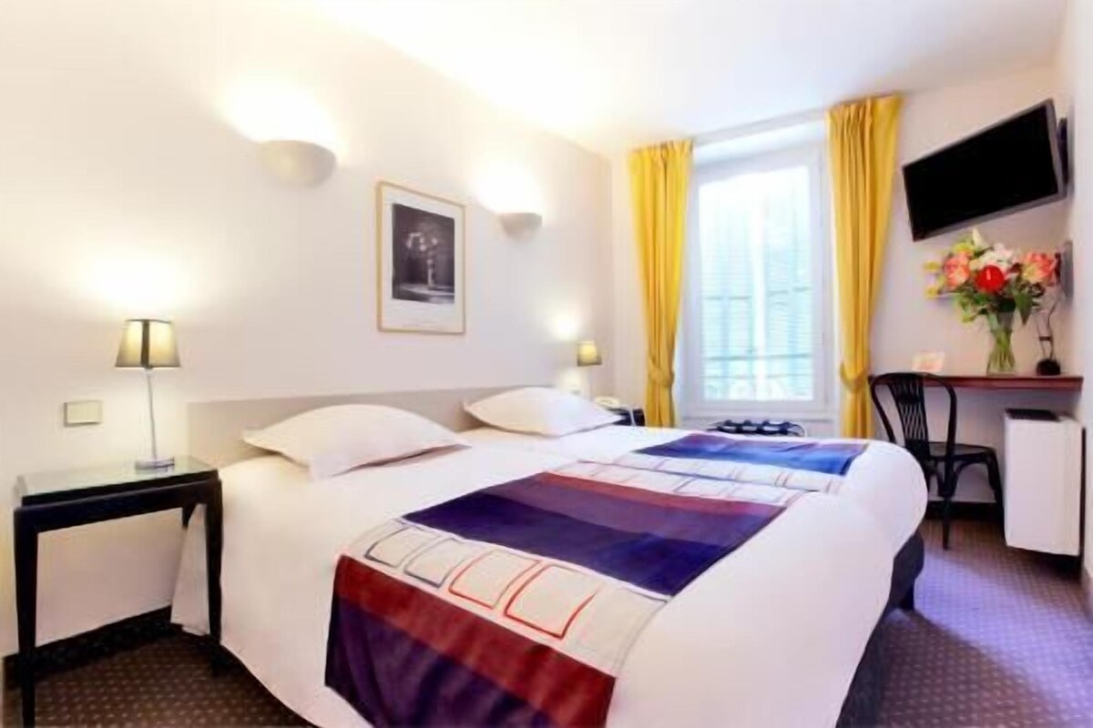 Habitación de hotel con cama, mesitas de noche, escritorio, silla, televisor y ventana con cortinas.