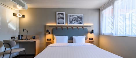Habitación de hotel con cama, escritorio con silla, ventana con cortinas translúcidas y dos cuadros en la pared.