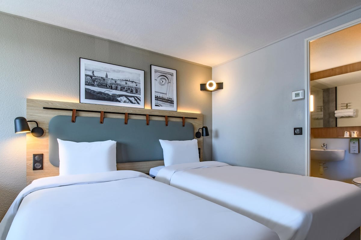 A hotel room with two beds, a wooden headboard, wall-mounted lamps, and framed pictures.