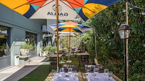 An outdoor dining area with tables and chairs under colorful umbrellas, surrounded by greenery and a building facade. - Campanile PRIME - Nîmes Centre (Nîmes)