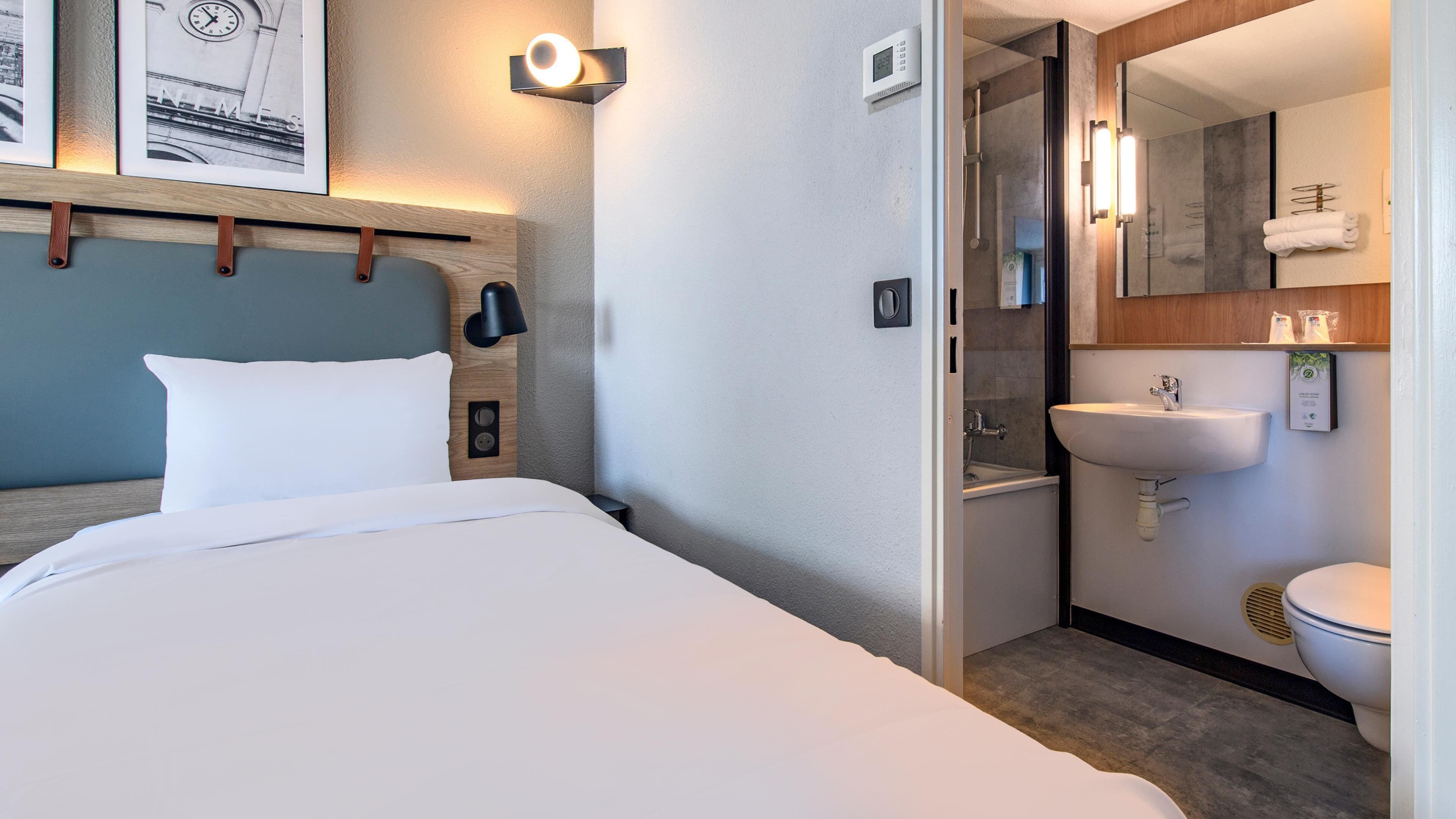 A hotel room with a bed, a bedside table, a wall-mounted clock, a light fixture, a shower area, a sink, and a toilet.