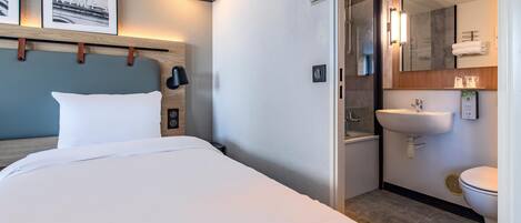 A hotel room with a bed, a bedside table, a wall-mounted clock, a light fixture, a shower area, a sink, and a toilet.