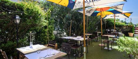 Un espacio de comedor al aire libre con mesas, sillas, sombrillas y follaje.