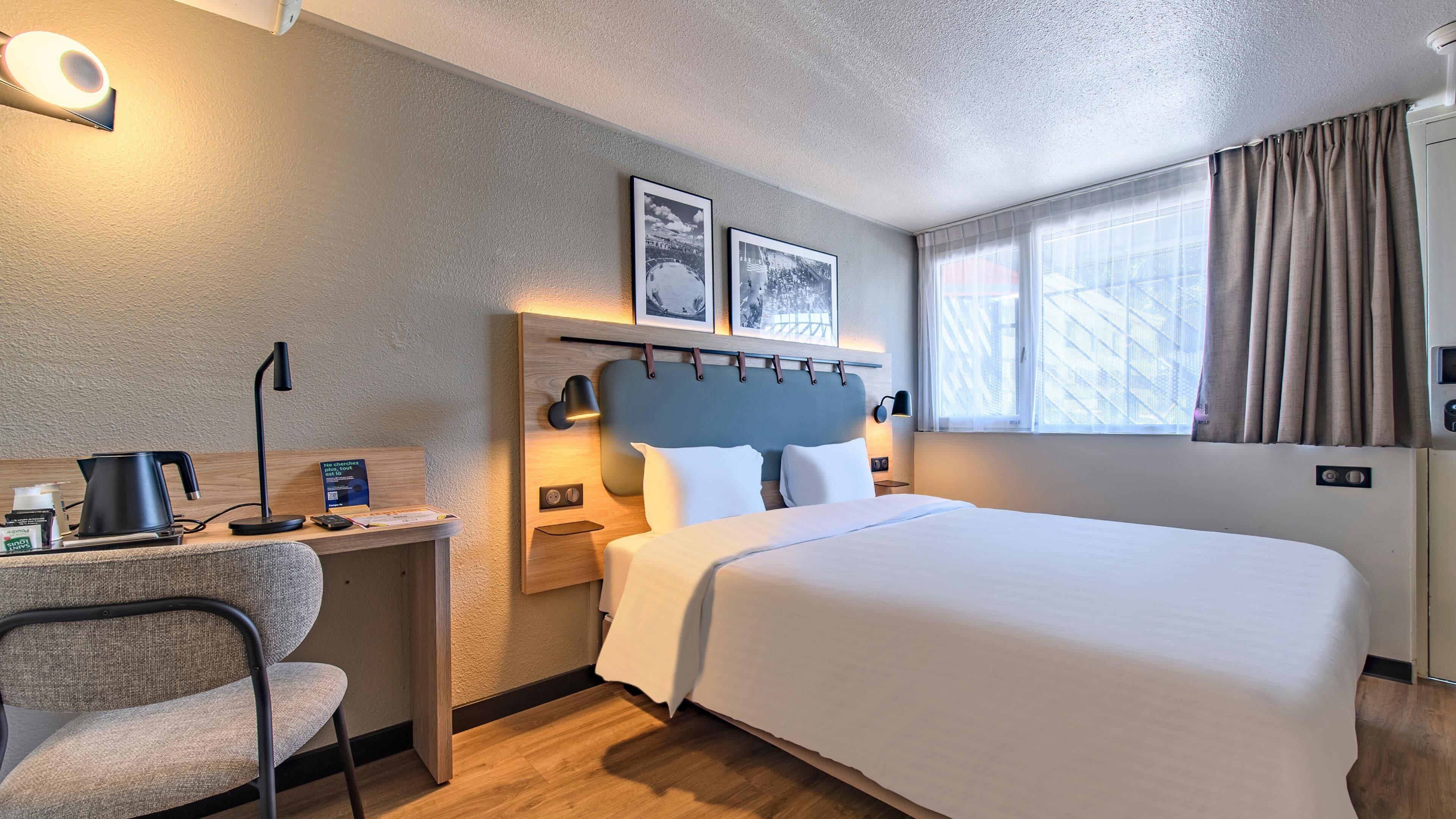 A hotel room with a large bed, a desk with a chair, a coffee maker, and a nightstand with a lamp.