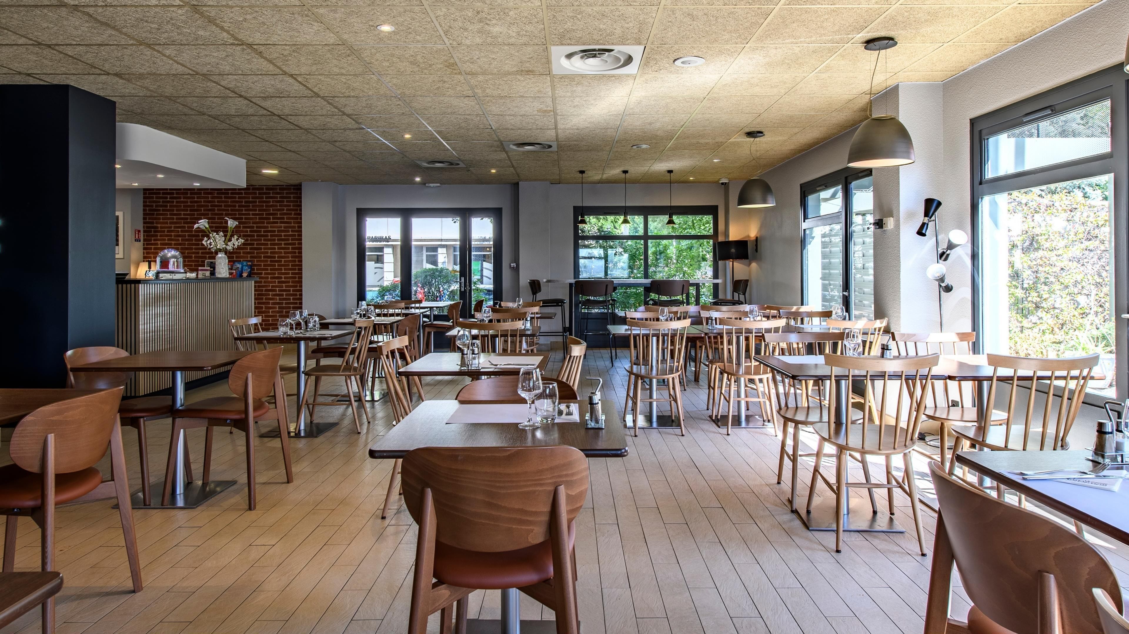 Un restaurante espacioso con mesas y sillas de madera, ventanas amplias con vistas a zonas verdes y un diseño de techo moderno.
