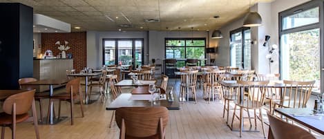 Un restaurante espacioso con mesas y sillas de madera, ventanas amplias con vistas a zonas verdes y un diseño de techo moderno.