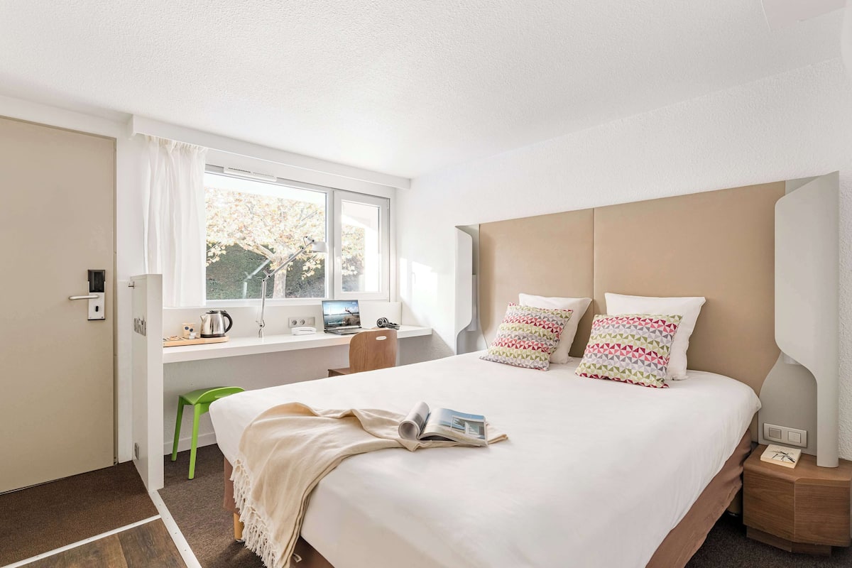 A modern hotel room with a large bed, a desk with a laptop, and a window with a view of trees.