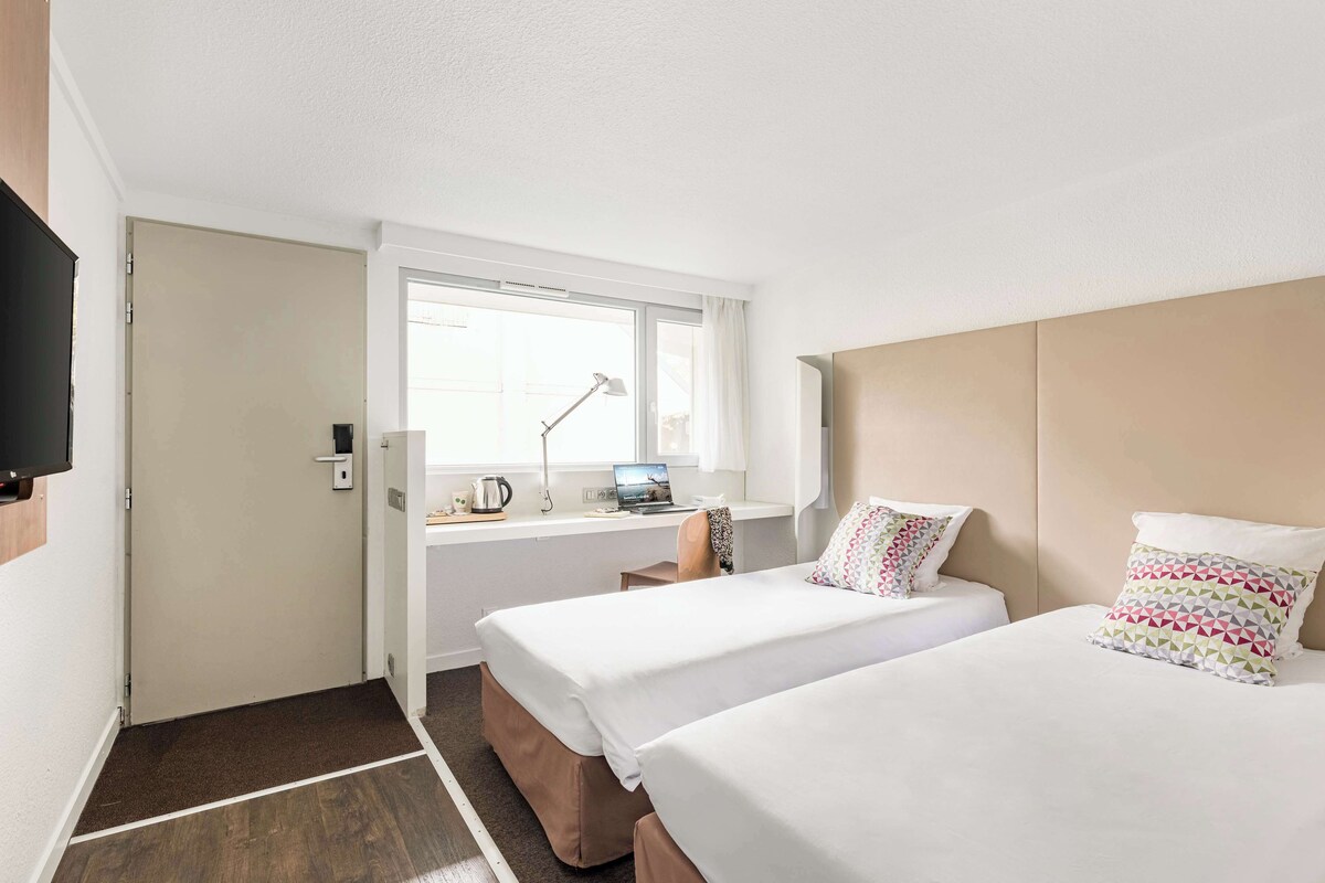 Habitación de hotel con dos camas, un escritorio con una computadora portátil, una ventana y un televisor en la pared.