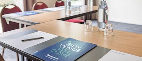 Een vergaderruimte met een tafel gedekt voor een vergadering, inclusief een 'Bien Venue Well Come' welkomstkaart, een fles, glazen en een pen op tafel.