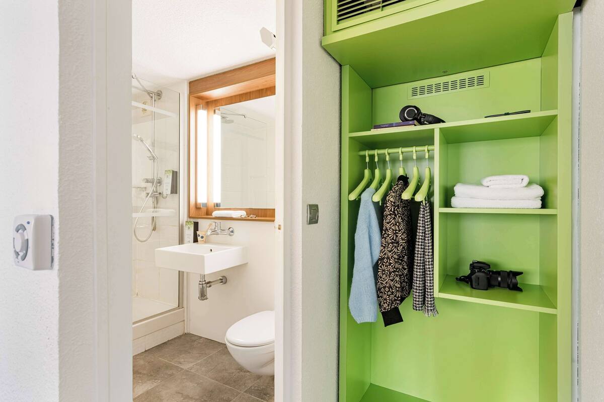 A bathroom with a shower, sink, and toilet. There is a green storage unit with hangers and towels.
