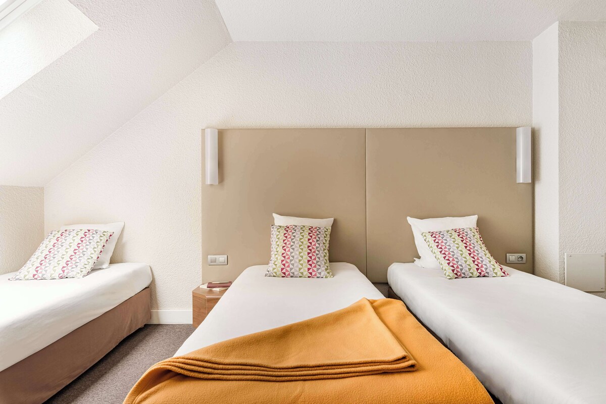 Una habitación de hotel con dos camas, cada una con una almohada estampada, un cabecero color beige y una manta marrón en una de las camas.