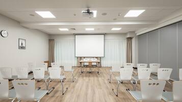 Une salle de conférence avec des chaises blanches, un écran de projection, une horloge et une fenêtre avec des rideaux.