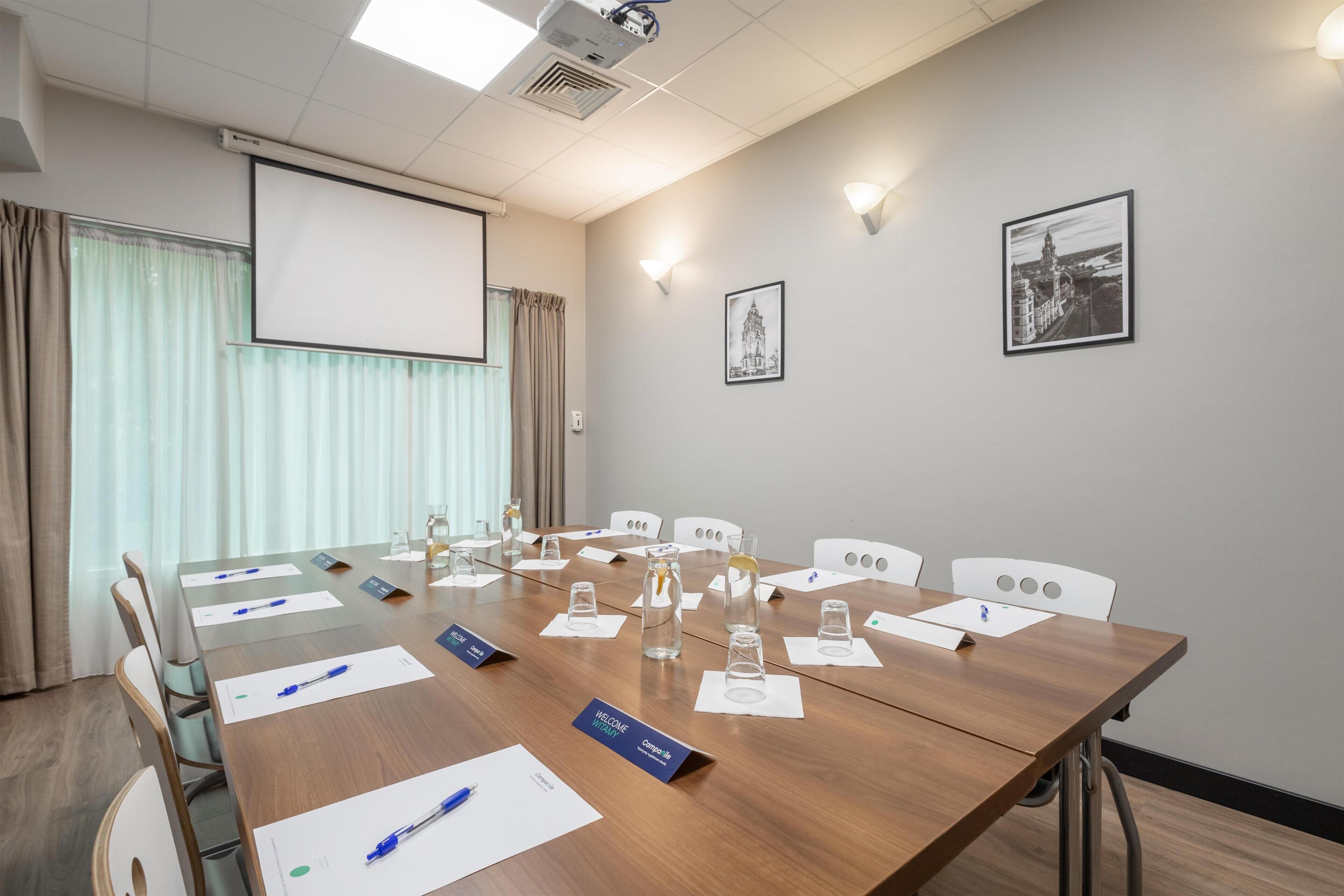 A conference room with a long table, chairs, nameplates, water glasses, and a projector screen.