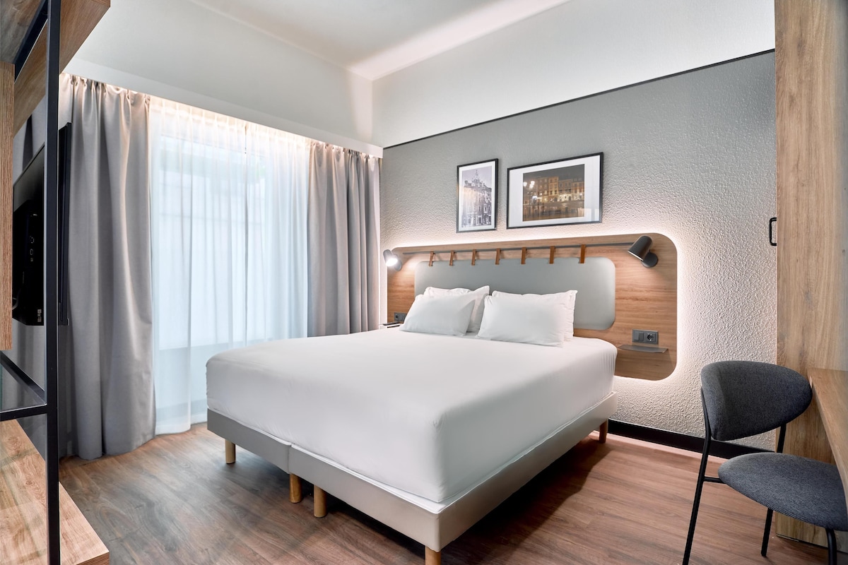 Une chambre d’hôtel moderne avec un grand lit, une télévision, une chaise et une table d’appoint en bois.