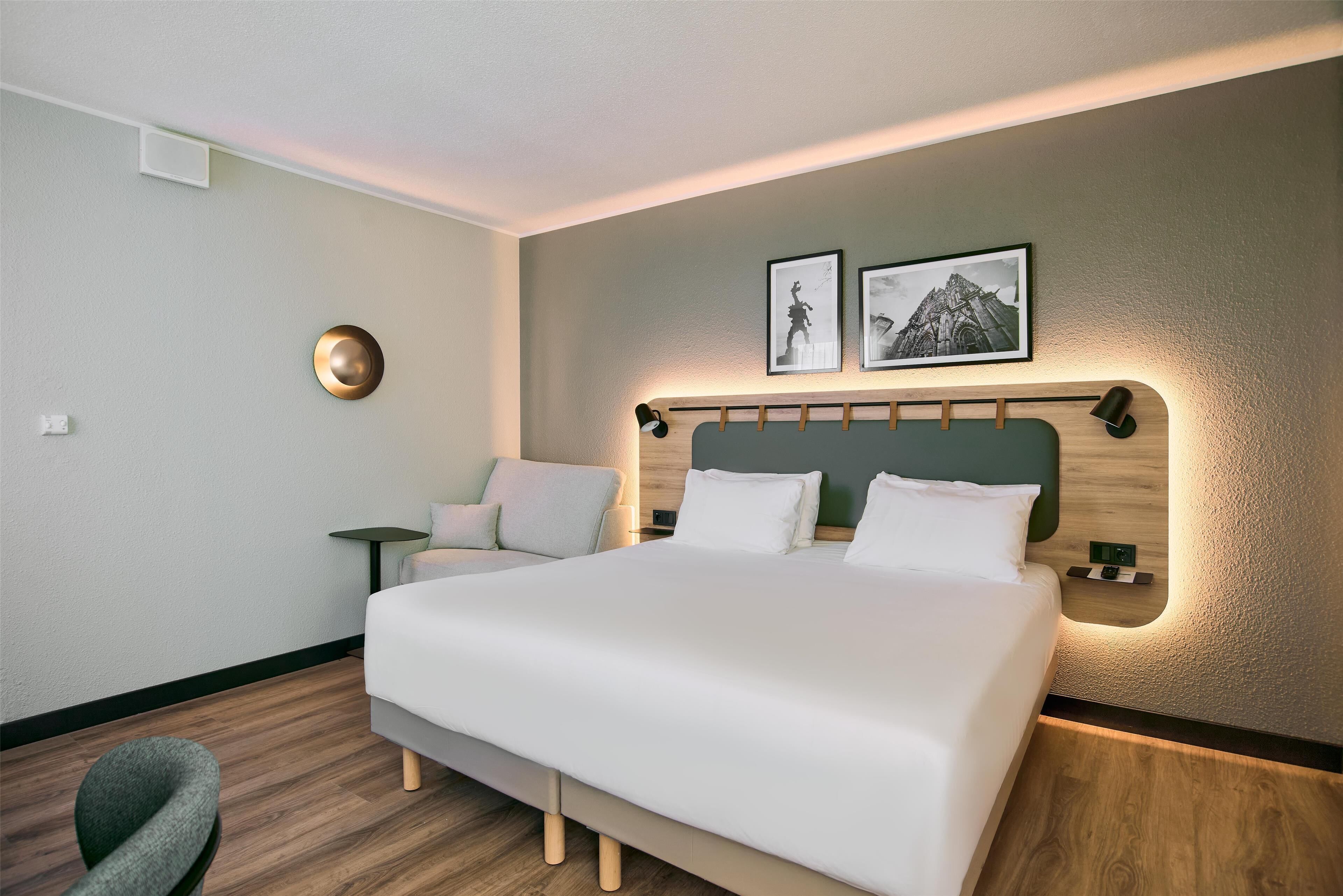 A modern hotel room with a large bed, a small side table, a chair, and two framed pictures on the wall.