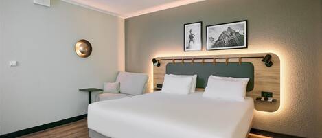 A modern hotel room with a large bed, a small side table, a chair, and two framed pictures on the wall.