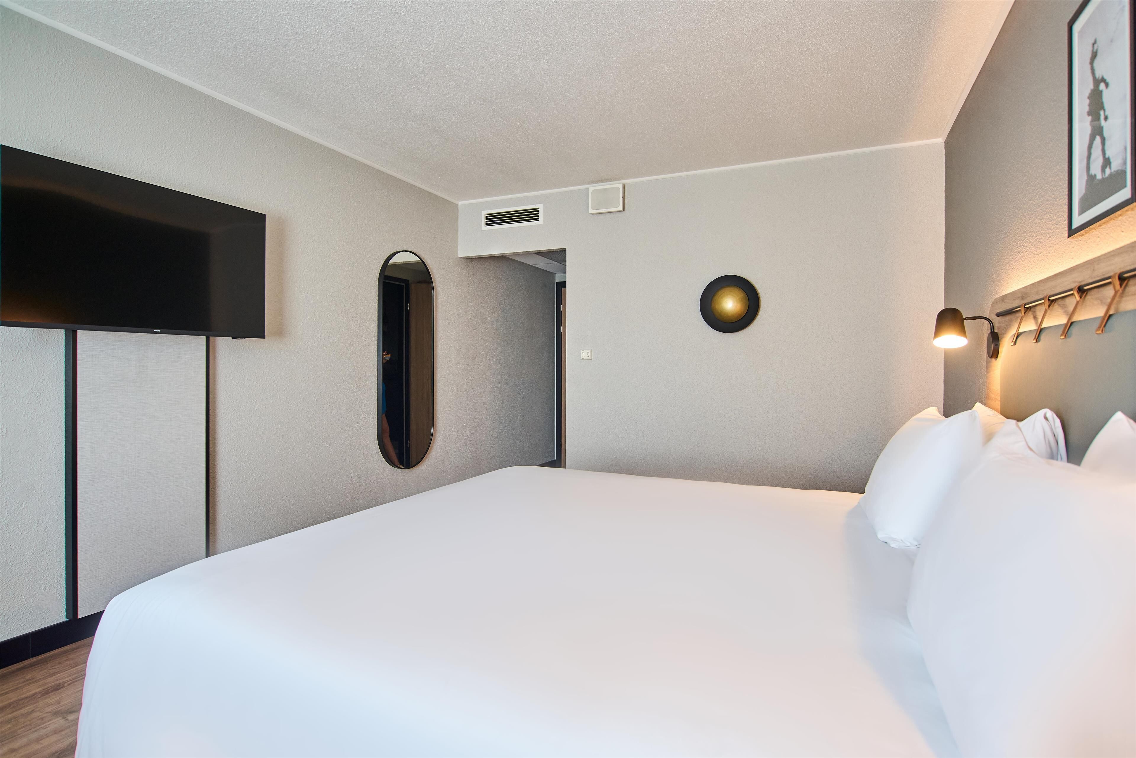 A hotel room with a large bed, a flat-screen TV mounted on the wall, a round wall clock, and a bedside lamp.
